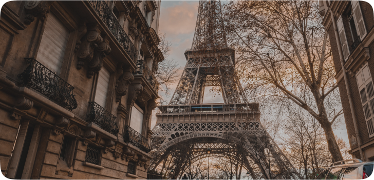 Imagem da Torre Eiffel, uma torre triangular feita de metal que está centralizada em uma famosa praça da França, vista de uma rua que o leva em direção a ela.